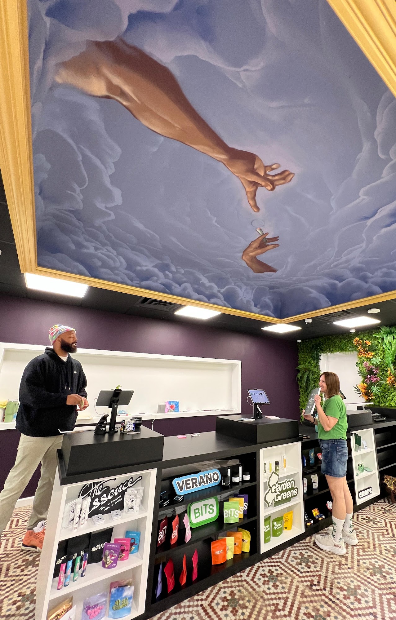 Hamm & Chaz budtender helping a customer at the counter beneath the ceiling mural — cannabis delivery to Bloomfield, NJ